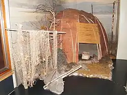 Летнее нанайское жилище из бересты и ивовых прутьев. На заднем плане — вешала́ для ю́колы, спереди — рыбацкие сети и деревянный якорь.Хабаровский краевой музей имени Н. И. Гродекова