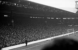 Photograph of a stand full of people. In front of the stand is a field and a goal.