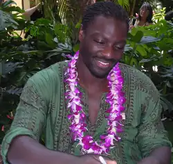 A black man, wearing a green shirt and a necklace of purple flowers.