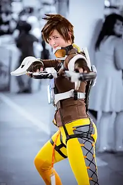 A young, Caucasian woman with short, spiky hair wearing a brown jacket and yellow leggings is facing the camera and extending two fake pistols; one in each hand.