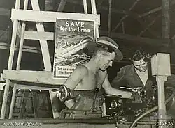 Black-and-white photograph of two soldiers working with a lathe. A poster behind them depicts a ship with hospital markings sinking by the bow and is captioned with «SAVE for the brave» and «Let us avenge the Nurses».
