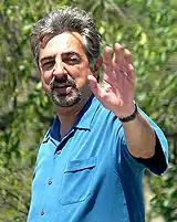 Colored photograph of a man, waving with his left arm. He is wearing a blue shirt.