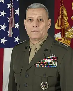 A color image of John Paxton, a white male in his Marine Corps Service A uniform. He is not wearing a hat, ribbons are visible as well as a basic parachutist badge. The Marine Corps flag and United States flag are visible in the background.