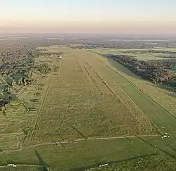 Поле с двумя травяными взлётно-посадочными полосами. По левой стороне (восточный край аэродрома) - заросшие травой капониры. Длинные тени от солнца. По краям - лес. Небо без облаков, с дымкой.