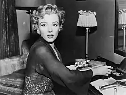  Monroe, wearing a transparent lace robe and diamond earrings, sitting at a dressing table and looking off-camera with a shocked expression