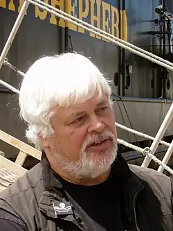 A heavyset man with white hair and a white beard, standing in front of a boat with the words «Shepherd» written on it in yellow paint