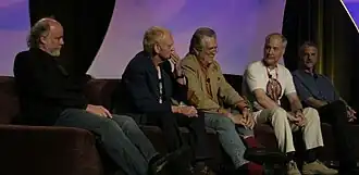 Пятеро пожилых мужчин сидят на диване, позади них — арочный декоративный фиолетовый фон. У Ралстона, откинувшегося на спинку сиденья, короткие волосы, он носит рубашку и брюки.