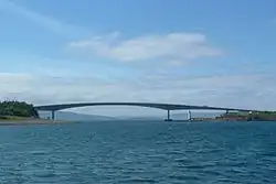  A body of blue water is spanned by a concave bridge of modern design in the middle distance. A small lighthouse can be seen beyond the bridge under its span.