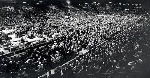 thousands of spectators in a large arena look towards a man standing behind a podium on a platform
